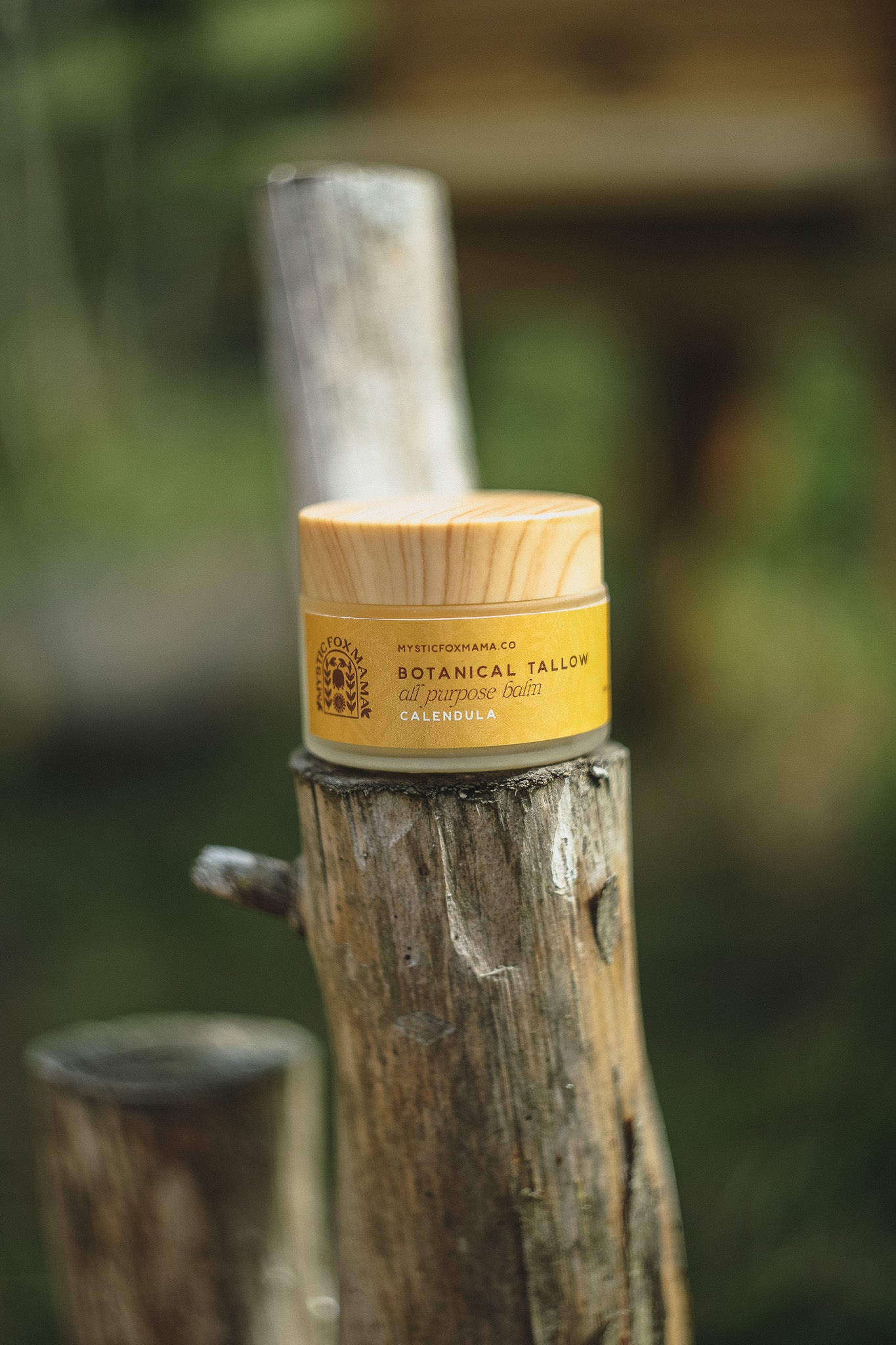 A jar of Botanical Tallow body butter with a bamboo lid and yellow label, resting on a rustic wooden post against a blurred outdoor green background.