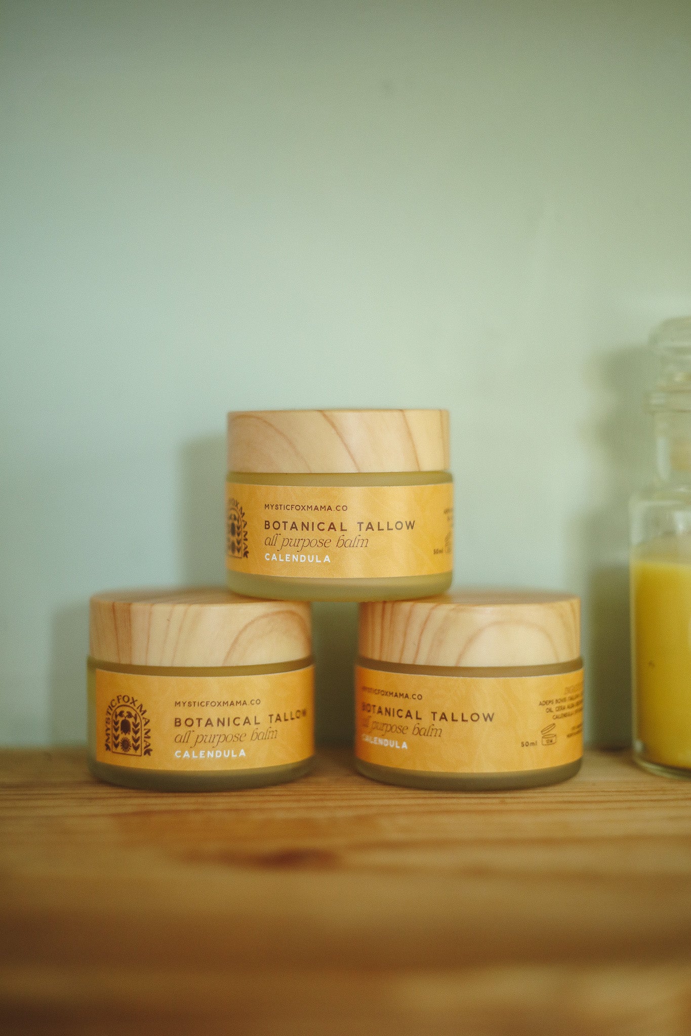 Three small jars of Botanical Tallow body butter arranged in a pyramid on a wooden surface against a pale green wall, with a glass bottle partially visible on the right.