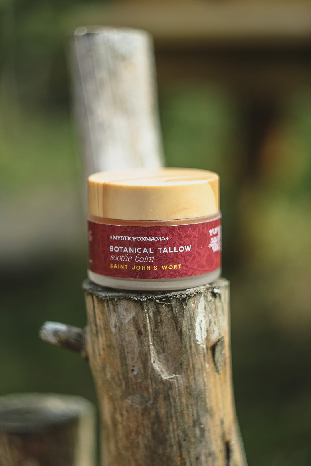 A jar of Harber's Churnham Botanical Tallow Hair Oil sits on a rustic wooden post, with a blurred outdoor garden background.