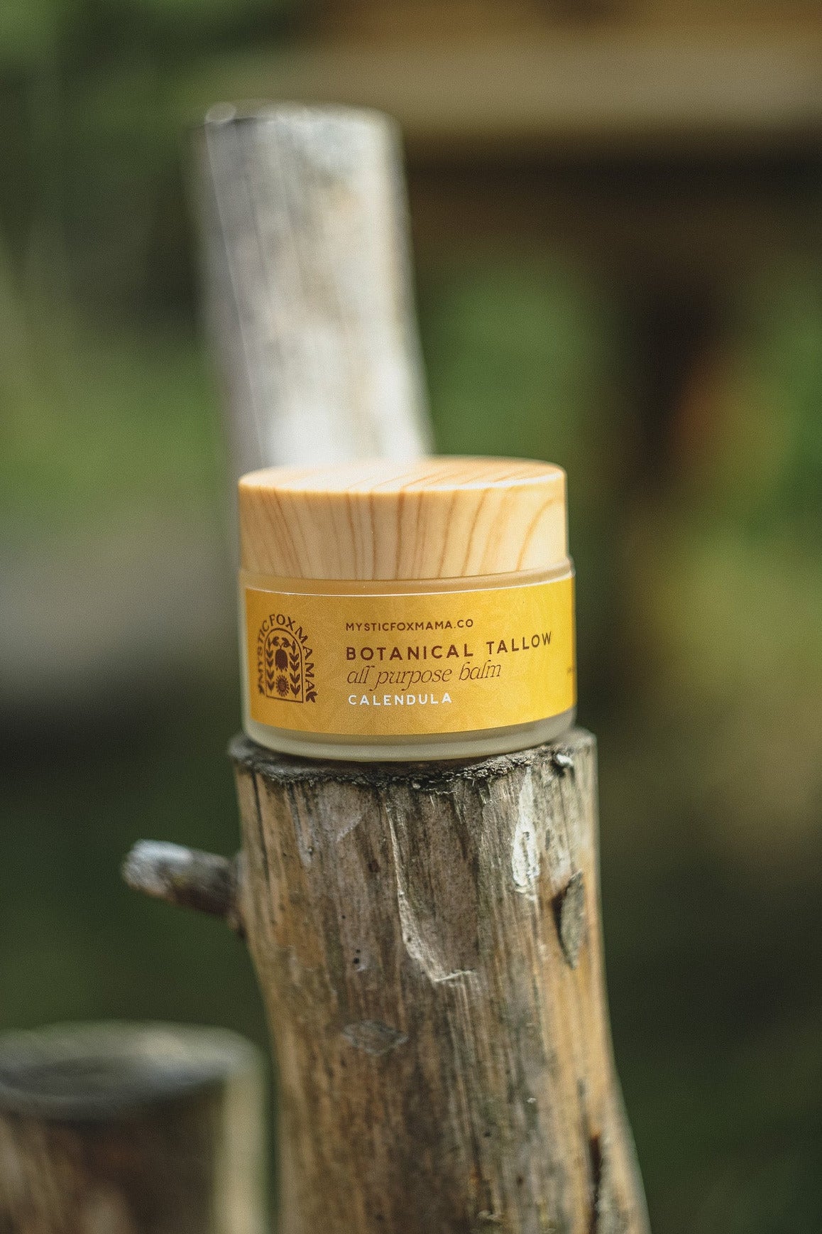 A small jar of Botanical Tallow body butter with a bamboo lid and yellow label, resting on a rustic wooden post with a blurred outdoor background.