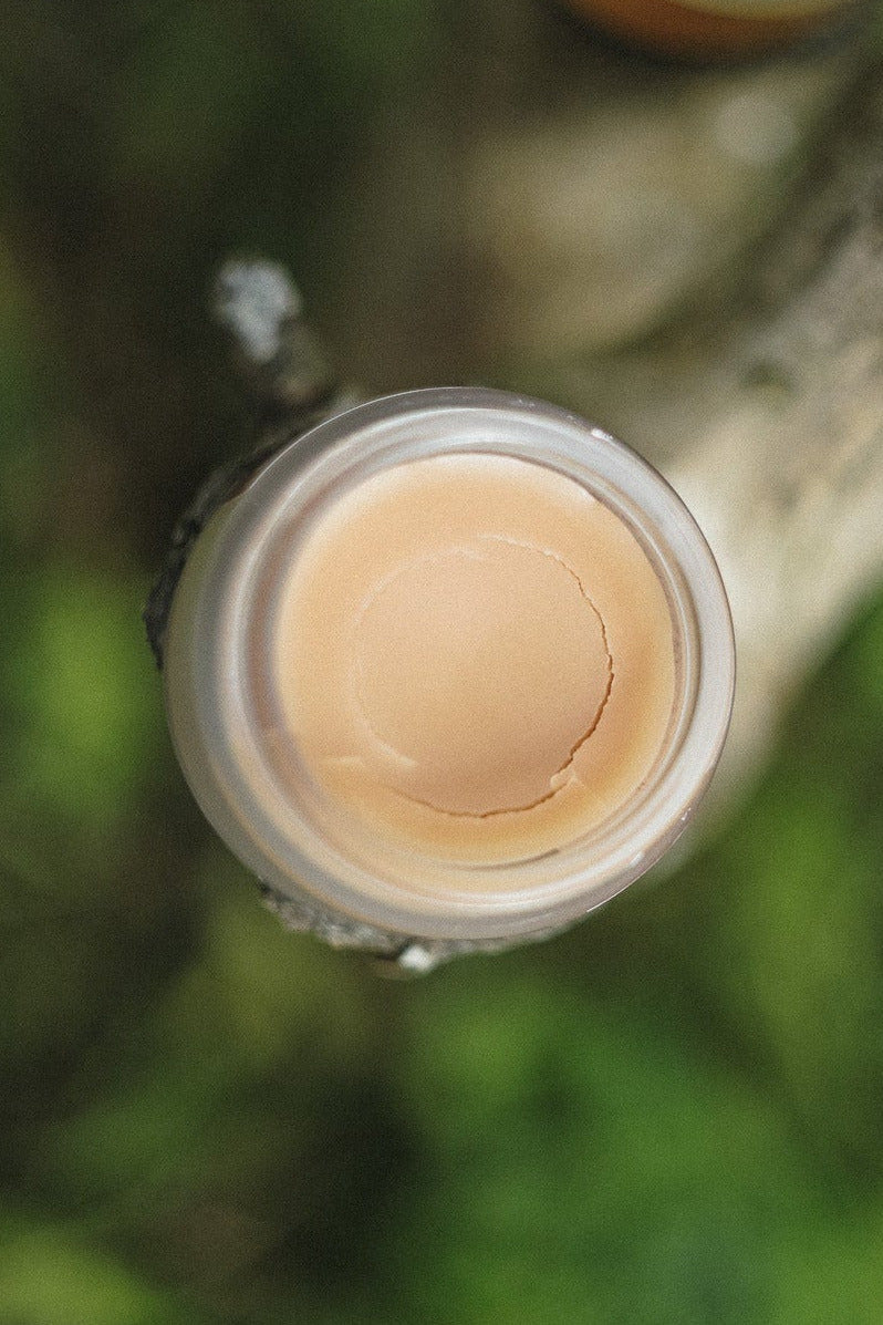 Top-down view of a clear jar filled with a pale peach-beige balm or cream, attached to a rustic wooden post outdoors, with another similar jar blurred in the background on green foliage.