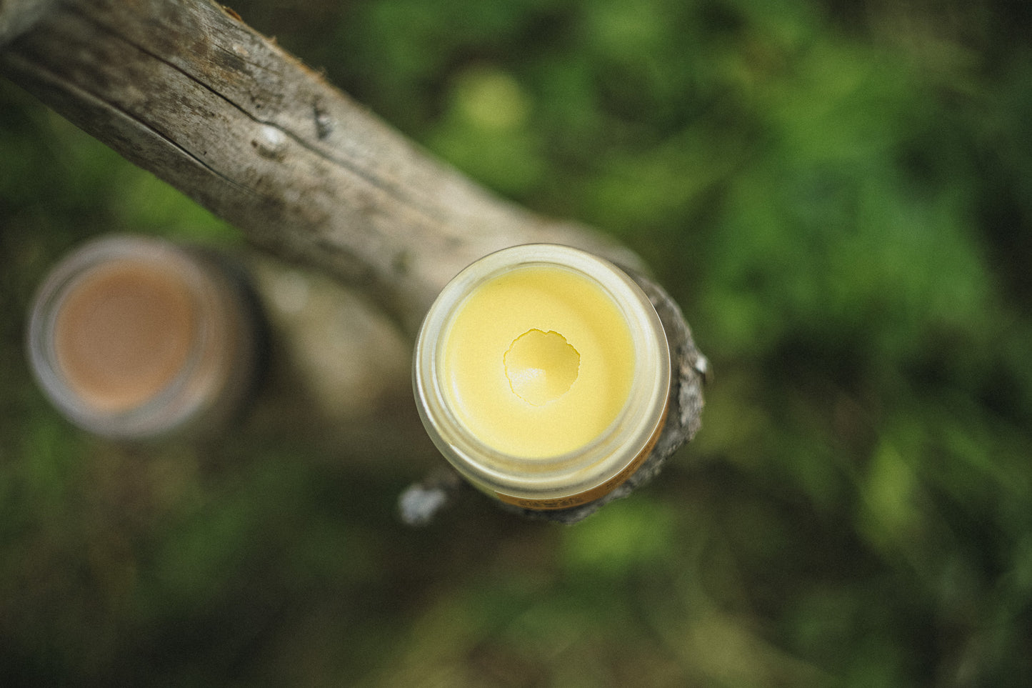 Top-down view of a small glass jar filled with pale yellow balm or wax, mounted on a weathered wooden post outdoors, with a second similar jar blurred in the background against a green natural backdrop.