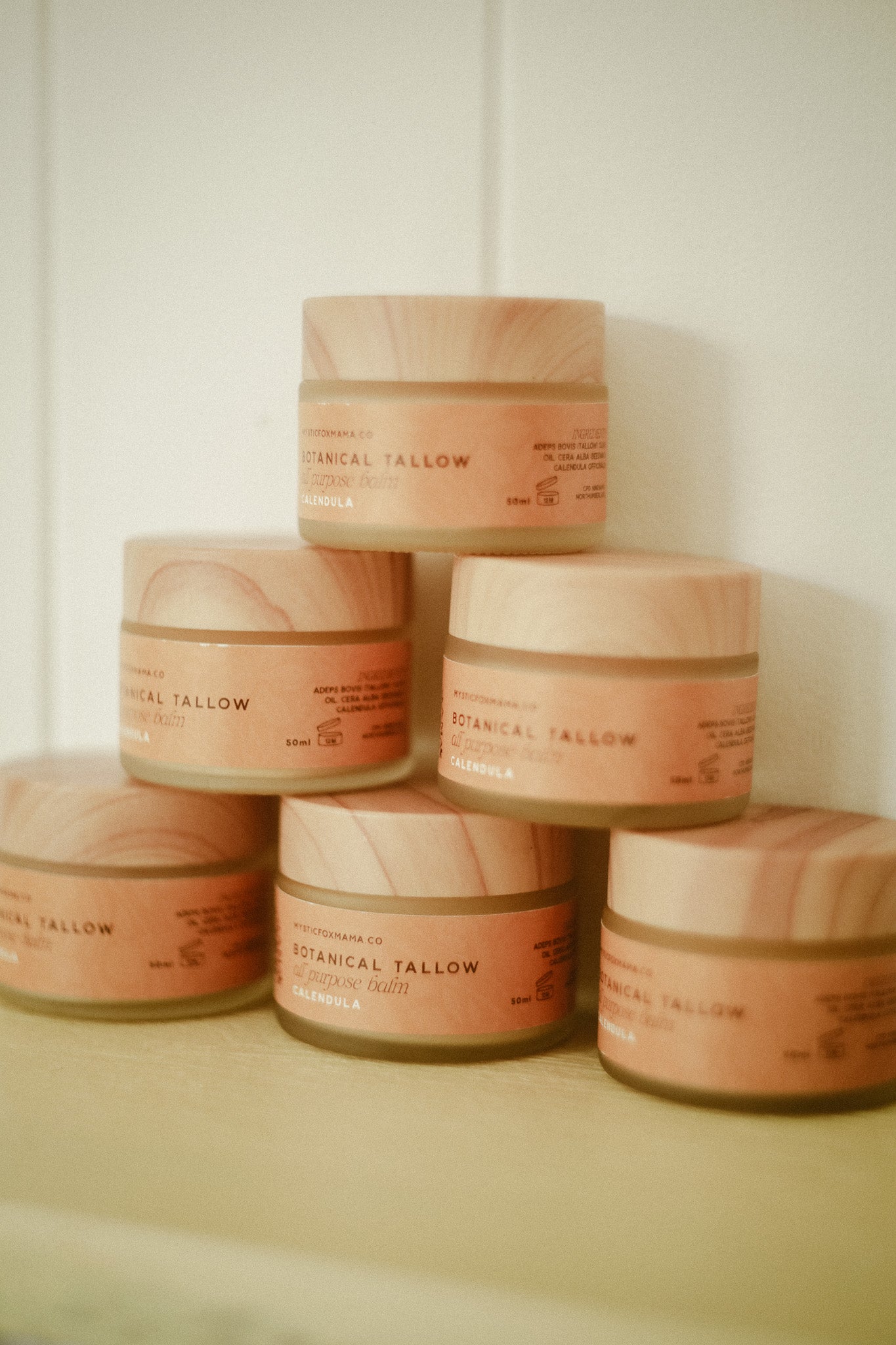 Five small cosmetic jars stacked in a pyramid arrangement on a light surface against a pale wall. The jars have light wood-grain lids and peach-colored labels with black text that reads 'BOTANICAL TALLOW'.