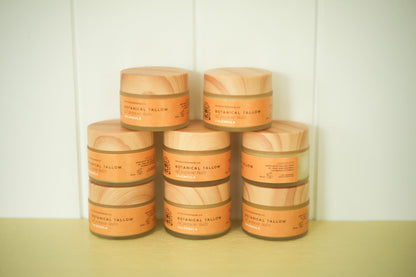 A stack of eight cosmetic jars with light wood-grain lids and orange labels, arranged in a pyramid on a pale yellow surface against a white paneled background.