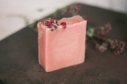 A rustic bar of soap with a piece partially cut out, revealing the pink interior, with rose petals and herbs on the top surface, placed on a dark, textured background.
