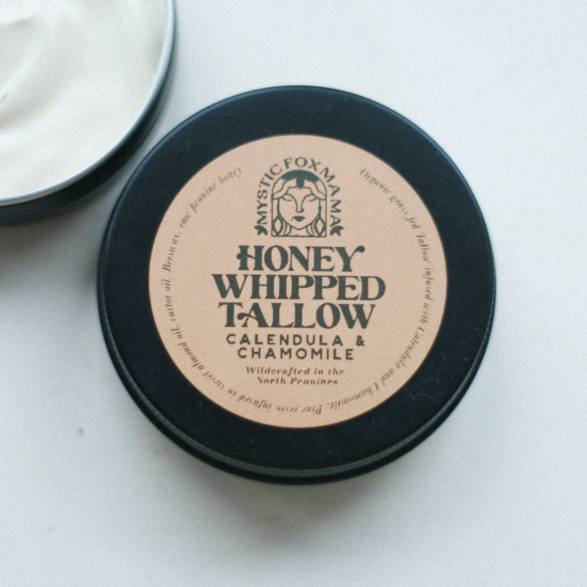 A small round container of Honey Whipped Tallow skin butter with a label that includes the product name and a brief description. The container is placed on a light surface, possibly a linen cloth.