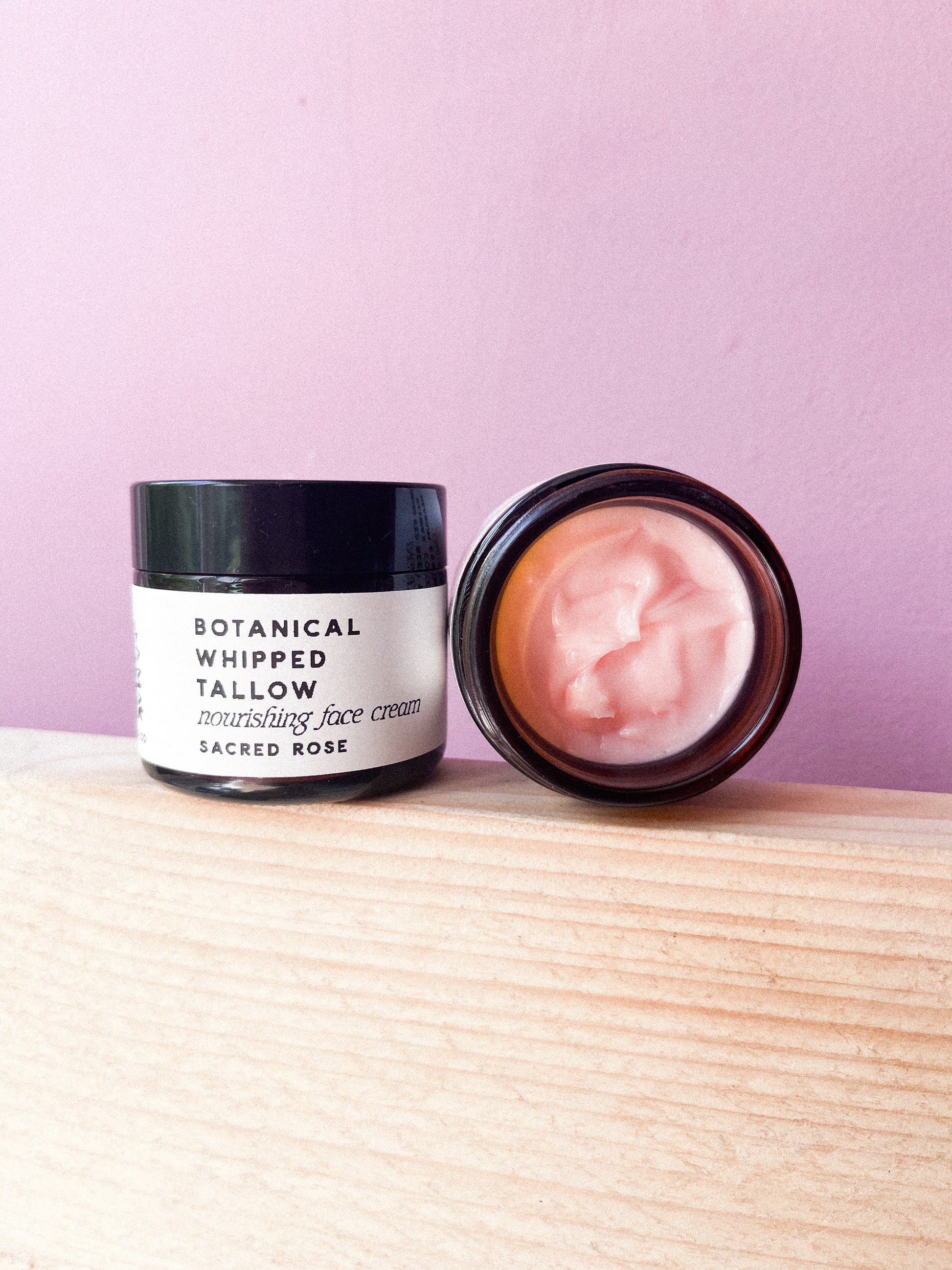 Two jars of Botanical Whipped Tallow face cream on a wooden surface with a pink background