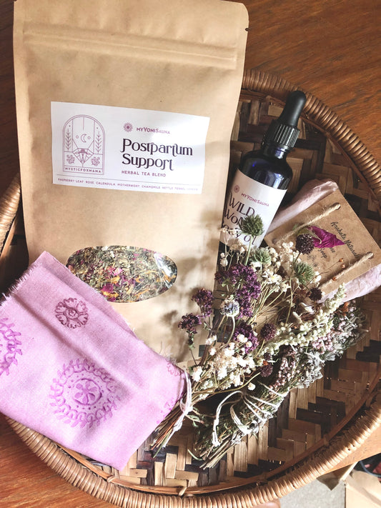 A basket containing various postpartum herbal support products including packages of herbs, bottles of oils, and a pink cloth.